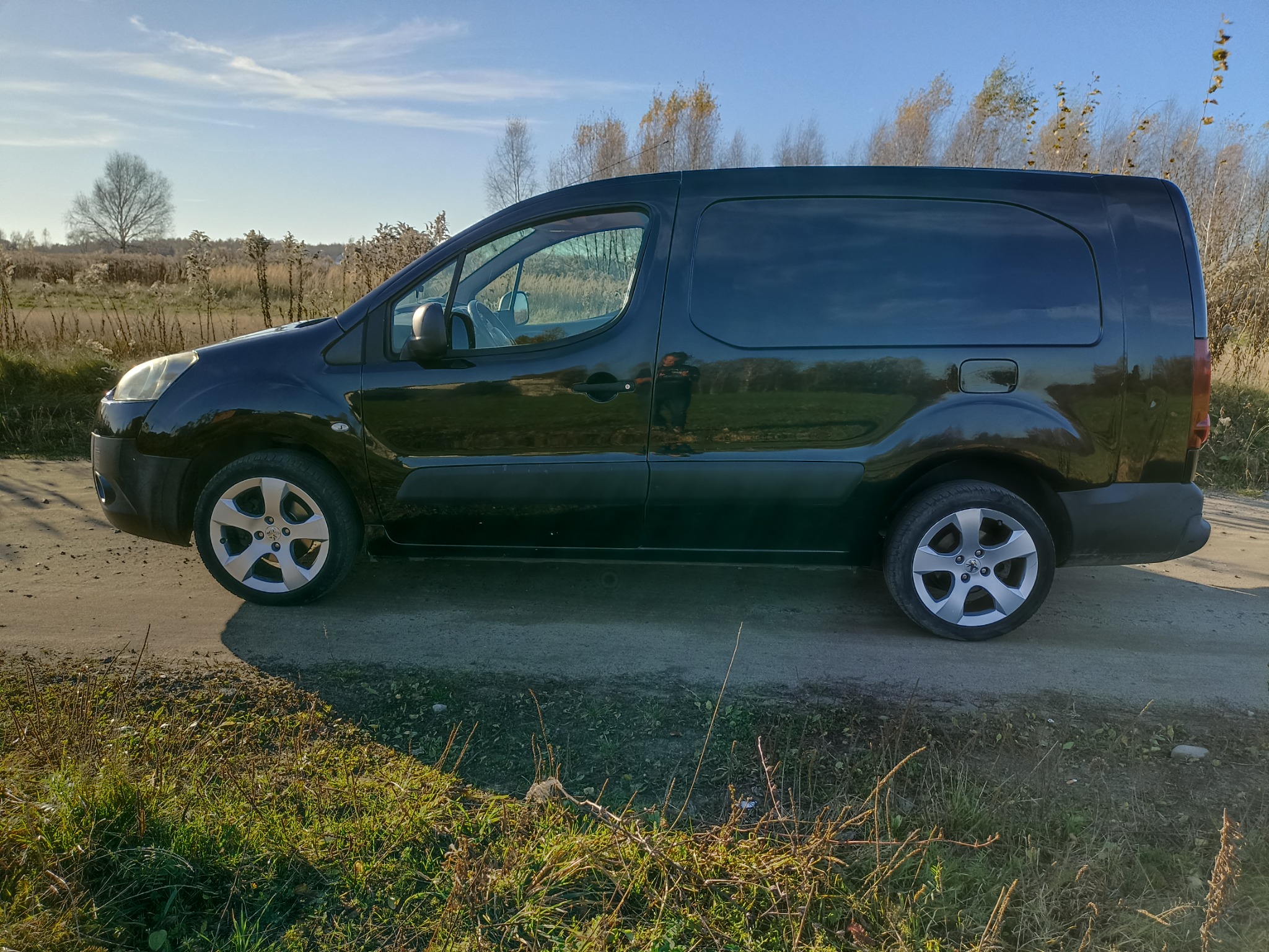 Czarny bus dostawczy Peugeot Partner zaparkowany na polnej drodze, jesienny krajobraz w tle.