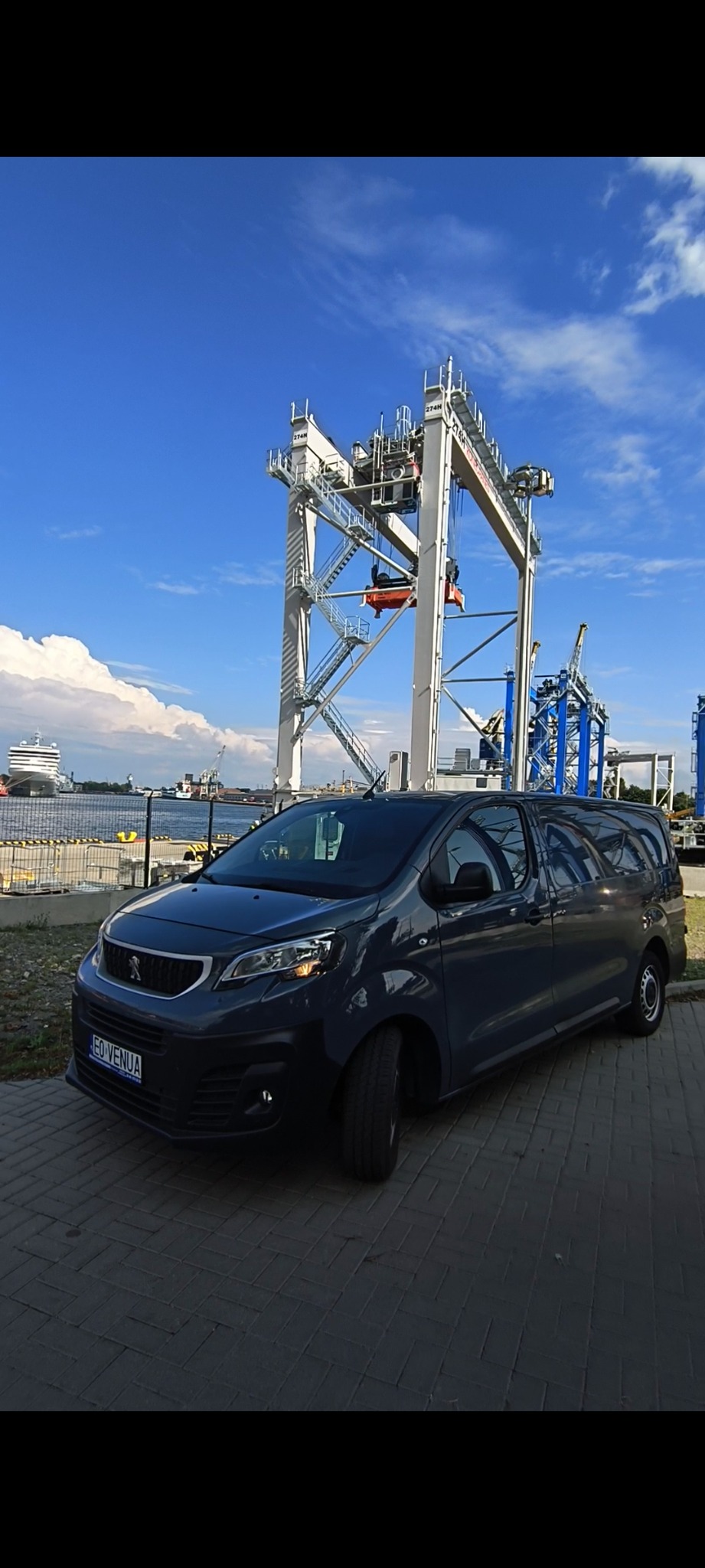 Szary bus Peugeot na tle portowego dźwigu kontenerowego i statku pasażerskiego, błękitne niebo z chmurami.