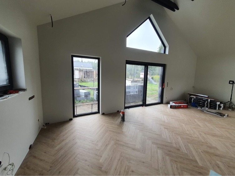 Wnętrze domu w trakcie wykańczania: jasne ściany, podłoga ułożona w jodełkę, duże okno trójkątne pod szczytem dachu, widoczne kable wystające ze ścian, okna z czarnymi ramami.