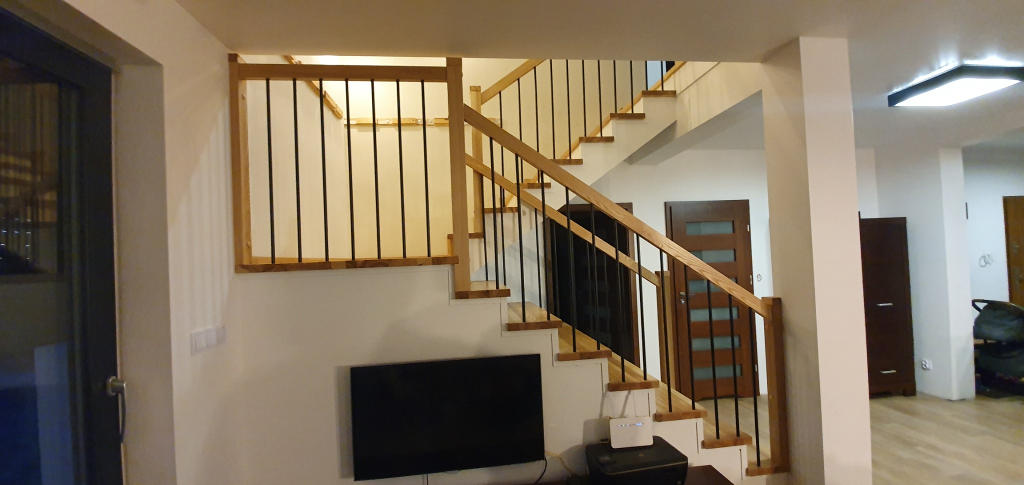 Drewniane schody z czarnymi balustradami w nowoczesnym domu, widok na telewizor i router pod schodami.