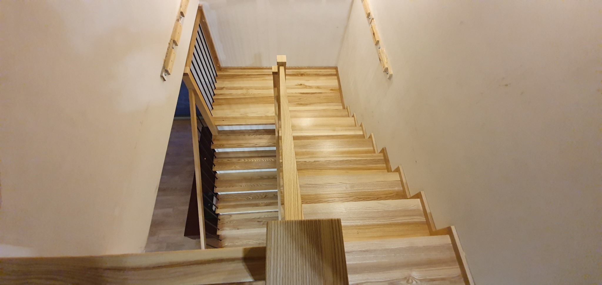 Widok z góry na jasne, drewniane schody z czarną metalową balustradą, zamontowane na beżowych ścianach w trakcie montażu, widoczne elementy mocujące na ścianie.