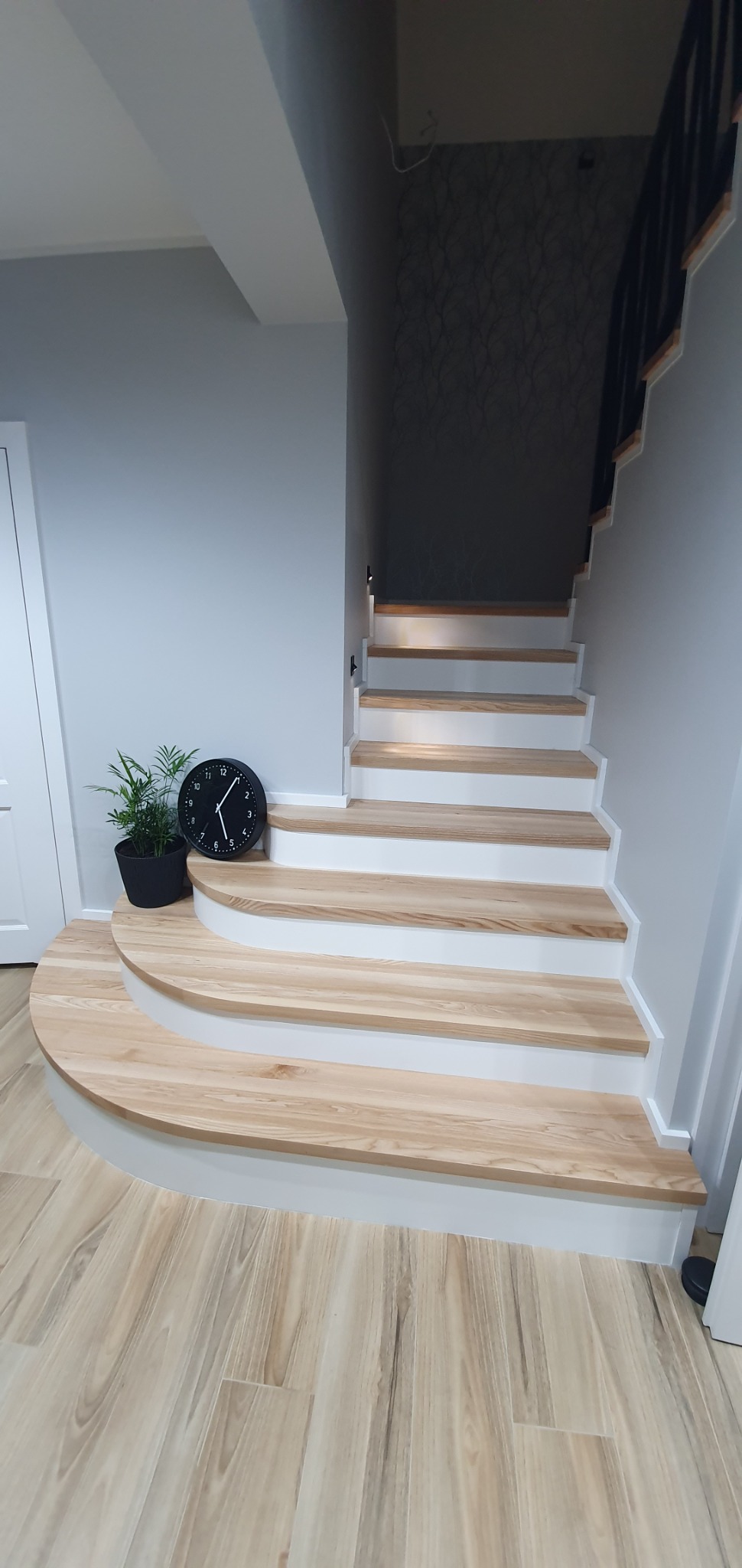 Drewniane schody z białymi podstopnicami, zaokrąglony pierwszy stopień, zegar i roślina doniczkowa obok schodów.