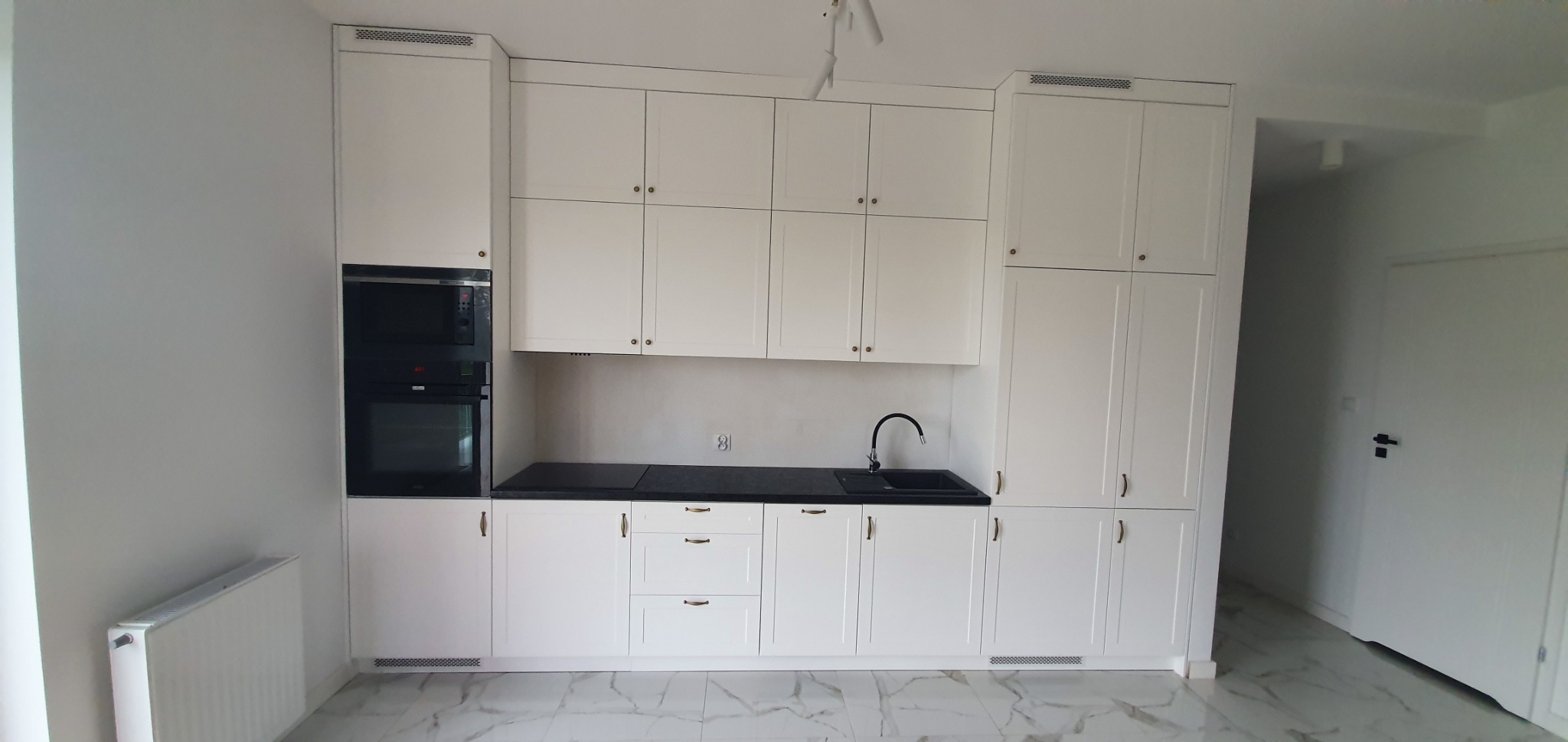 Biała zabudowa kuchenna z czarnym blatem, zintegrowanym piekarnikiem i mikrofalówką, oraz złotymi uchwytami na tle marmurowej podłogi.