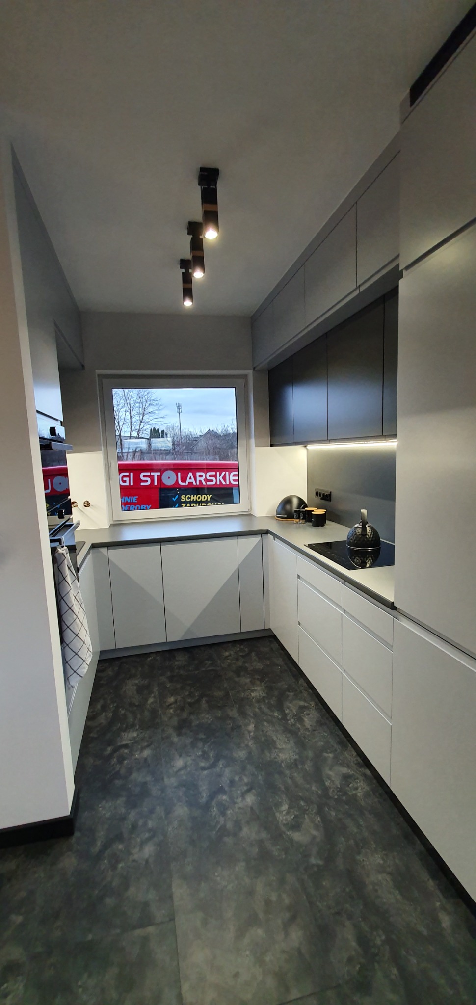 Nowoczesna kuchnia z szarymi i białymi szafkami, czarnym blatem, oświetleniem punktowym i widokiem z okna na zabudowania.