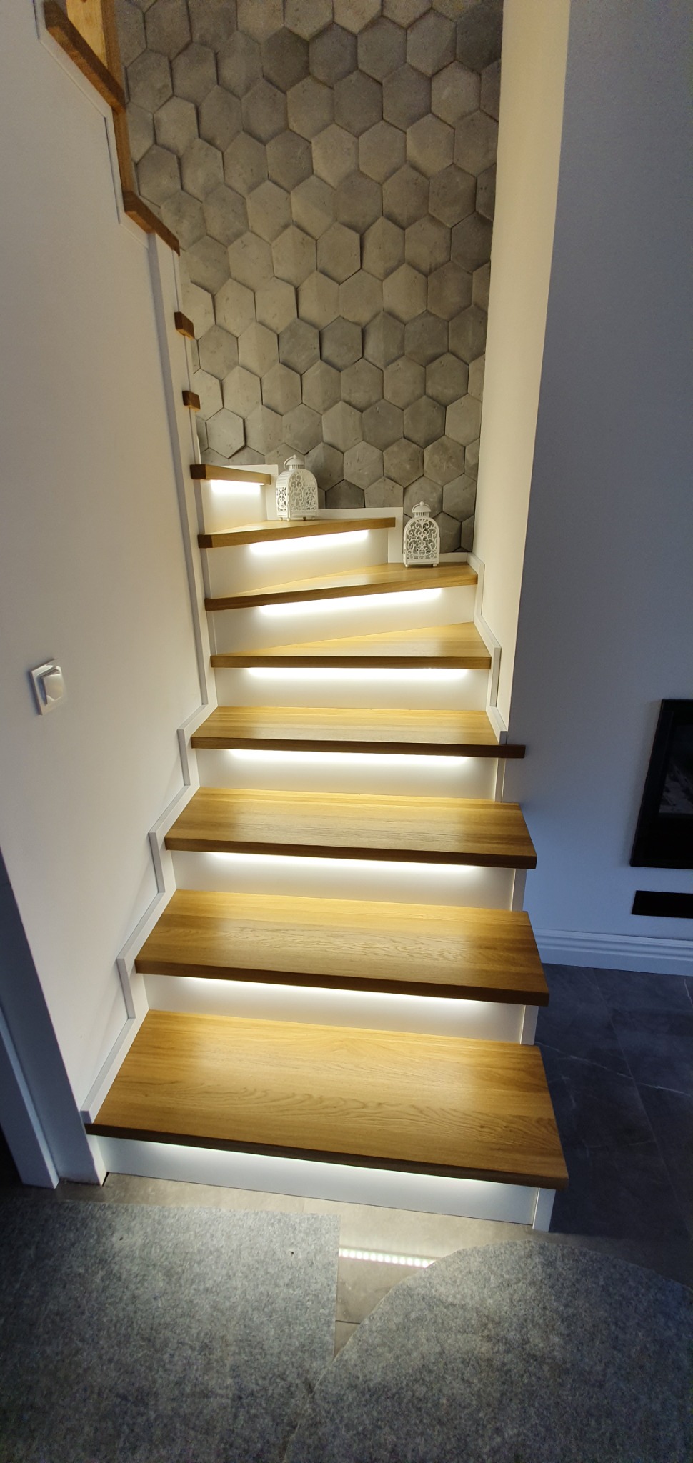 Nowoczesne schody drewniane z białymi bokami i podświetleniem LED, dekoracyjne lampiony i ściana z paneli 3D w kształcie plastra miodu.