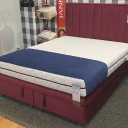 Tapicerowane łóżko kontynentalne w odcieniach bordo i bieli z granatowym bieżnikiem, widoczne pionowe przeszycia na wezgłowiu i metka producenta