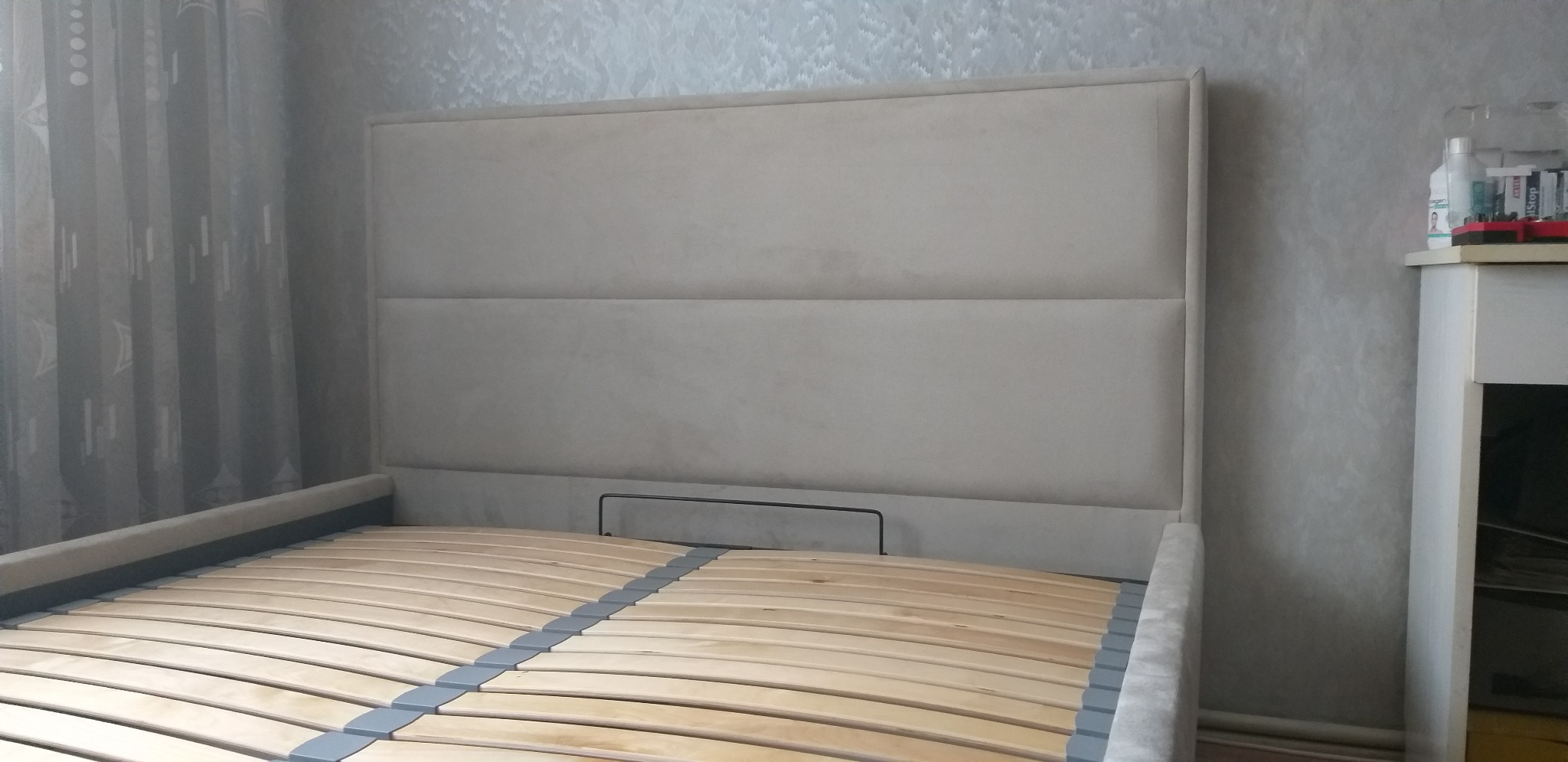 Szare łóżko z tapicerowanym wezgłowiem i drewnianym stelażem listwowym, widoczne przy zasłoniętym oknie i ścianie o fakturze betonu.