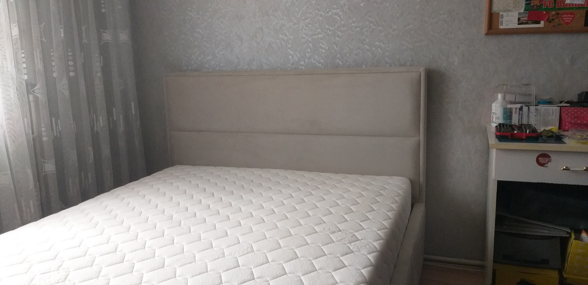 Beżowe łóżko tapicerowane z wysokim zagłówkiem w sypialni z wzorzystą tapetą i zasłonami, obok stolik nocny z przedmiotami.