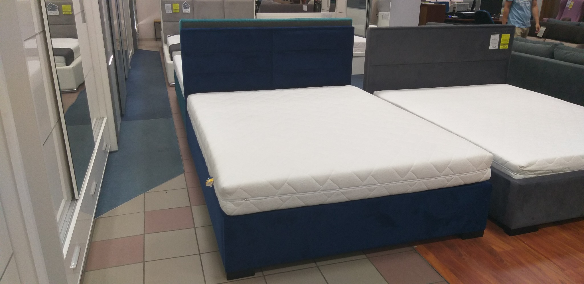 Niebieskie tapicerowane łóżko z pikowanym zagłówkiem i białym materacem w sklepie meblowym, obok szare łóżko z materacem.