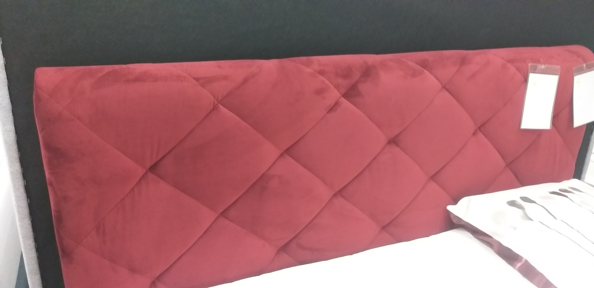 Czerwone, tapicerowane wezgłowie łóżka z pikowaniem w romby, widoczne metki i fragment poduszki.