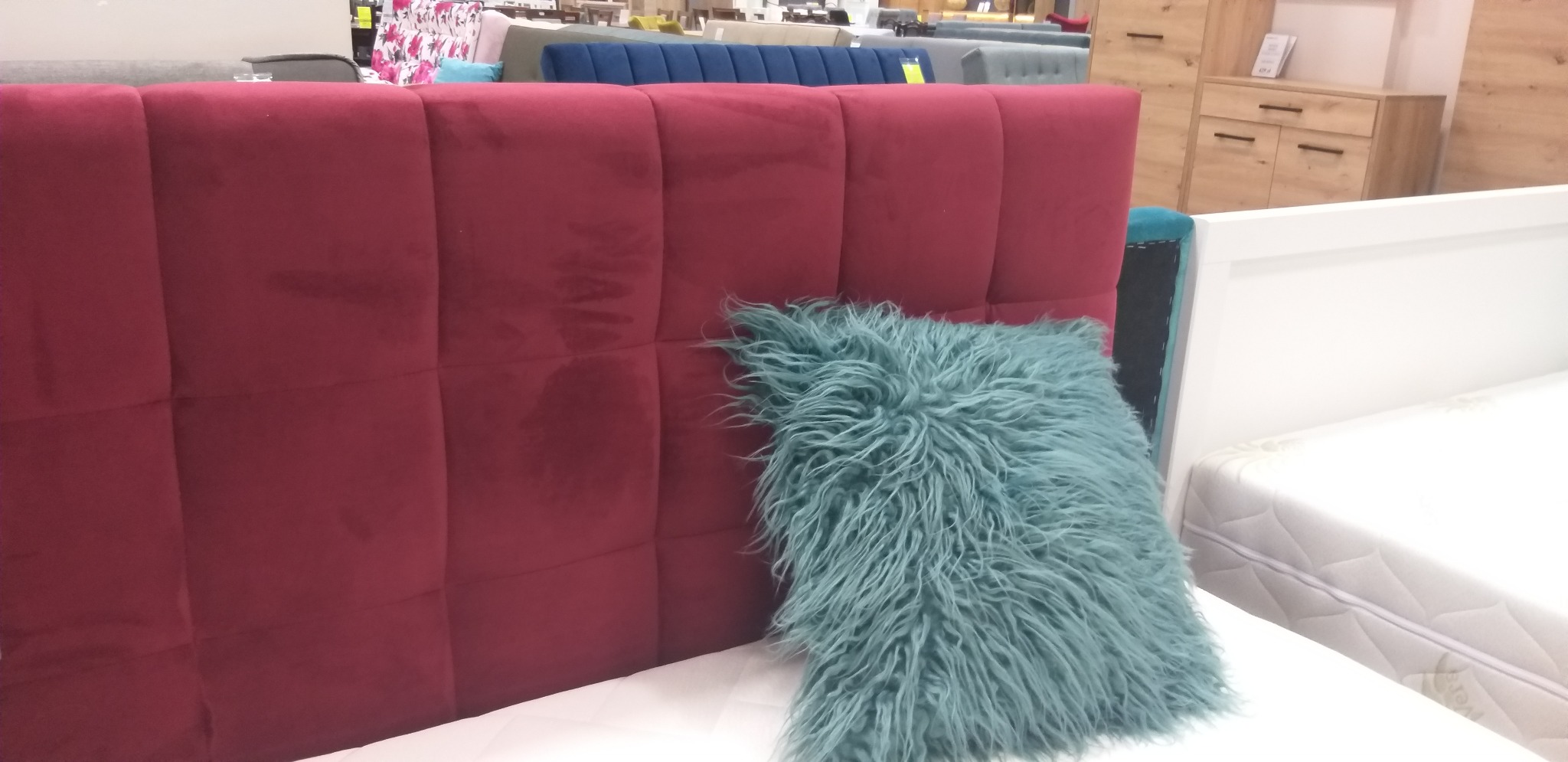 Czerwony, tapicerowany zagłówek łóżka z dekoracyjną, turkusową poduszką z długim włosiem, widok z boku na materac.