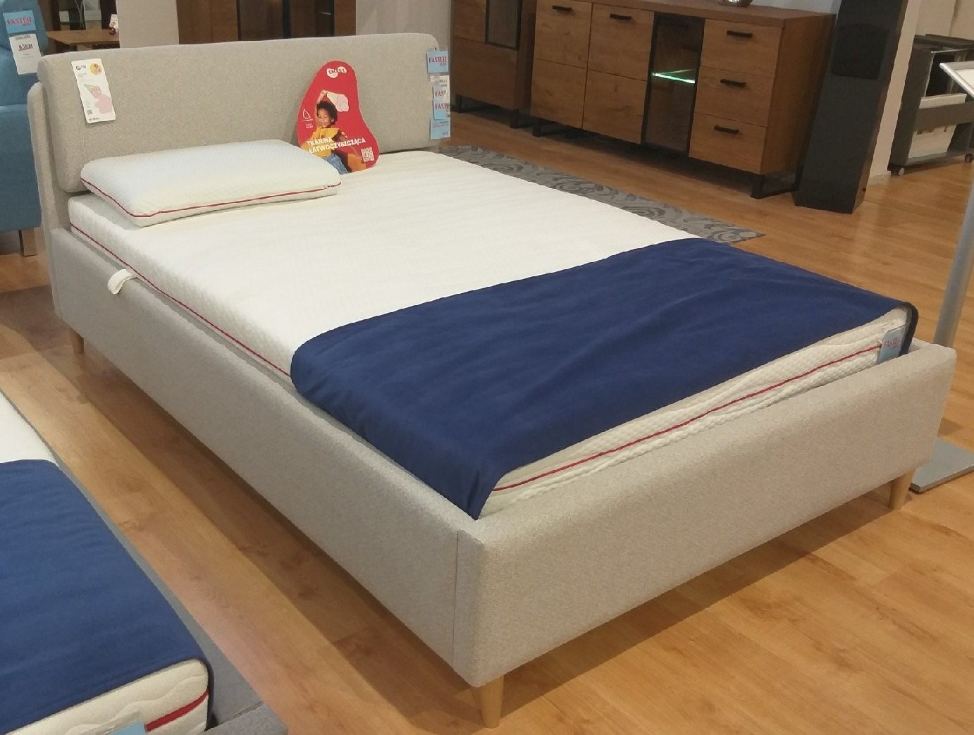 Szare łóżko tapicerowane z zagłówkiem, białym materacem z czerwoną lamówką i granatowym pledem, prezentowane w salonie meblowym.