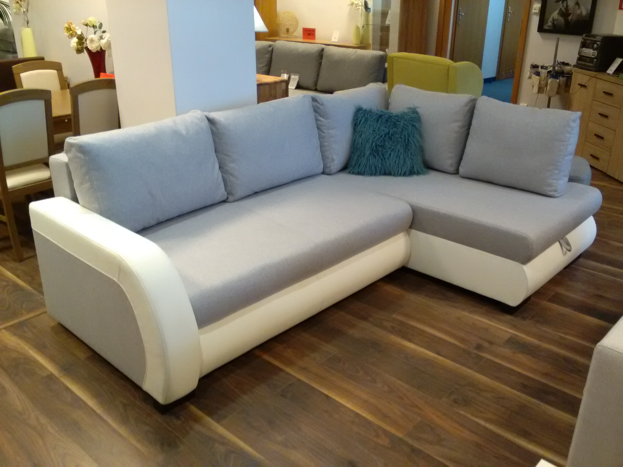 Szary narożnik z białym wykończeniem, ozdobiony turkusową poduszką, na drewnianej podłodze w salonie meblowym.