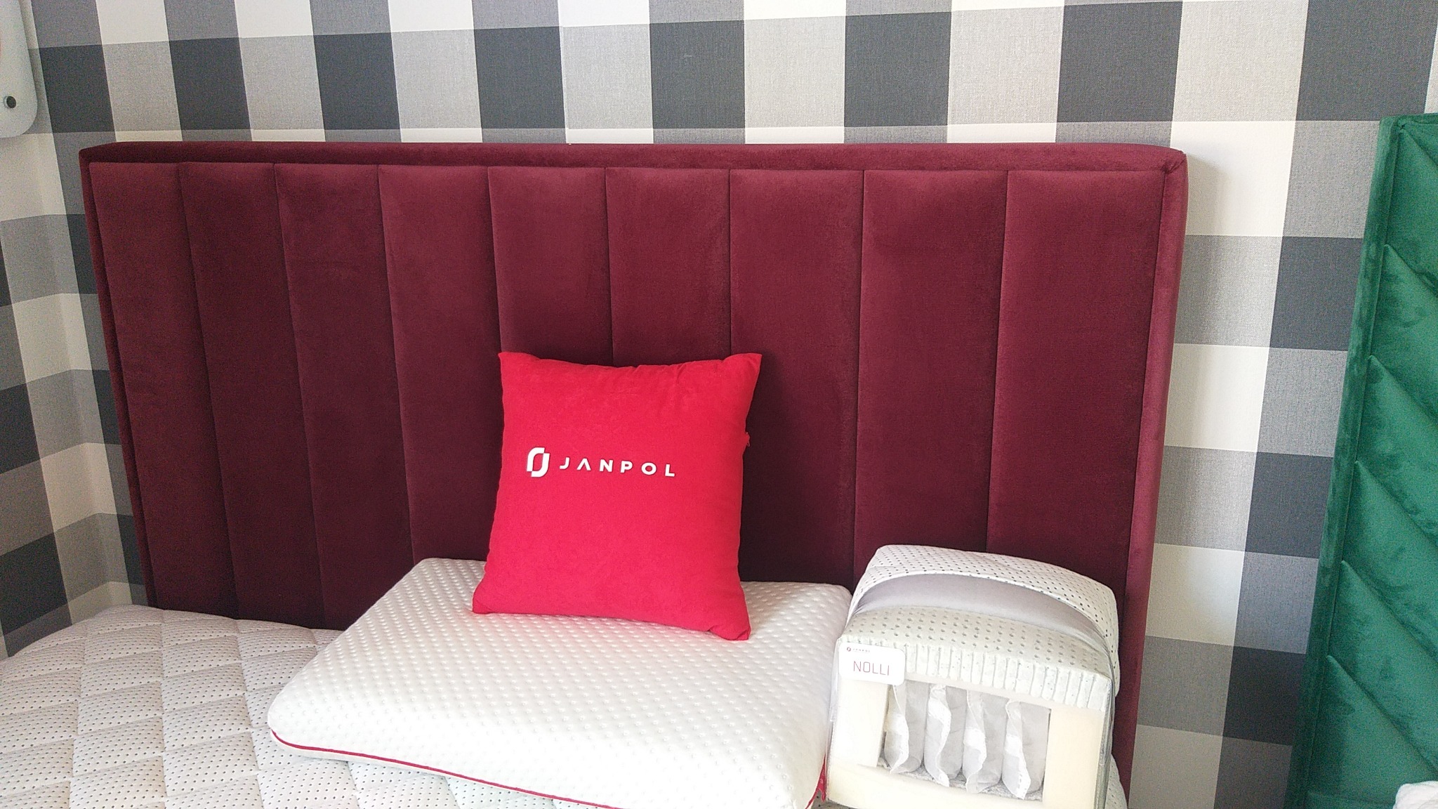 Czerwony, tapicerowany zagłówek łóżka z pionowymi przeszyciami, ozdobna poduszka z logo Janpol i materac z pianki memory, na tle ściany w szaro-białą kratę.