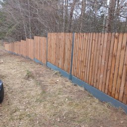 Ogrodzenia,montaż Mariusz Wawryńczuk - Długi, drewniany płot z szarym betonowym podmurówką, ciągnący się wzdłuż działki, otoczony drzewami bez liści. Widok z perspektywy ziemi.