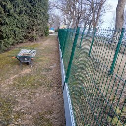 Ogrodzenia,montaż Mariusz Wawryńczuk - Zielone ogrodzenie panelowe na betonowej podmurówce, ciągnące się wzdłuż granicy działki z widoczną taczką i drzewami w tle. Ujęcie z perspektywy.