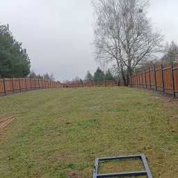 Ogrodzenia,montaż Mariusz Wawryńczuk - Długi, drewniany płot panelowy z metalowymi słupkami, otaczający trawnik z drzewami w tle. Widoczne elementy montażowe na pierwszym planie. Pochmurny dzień.