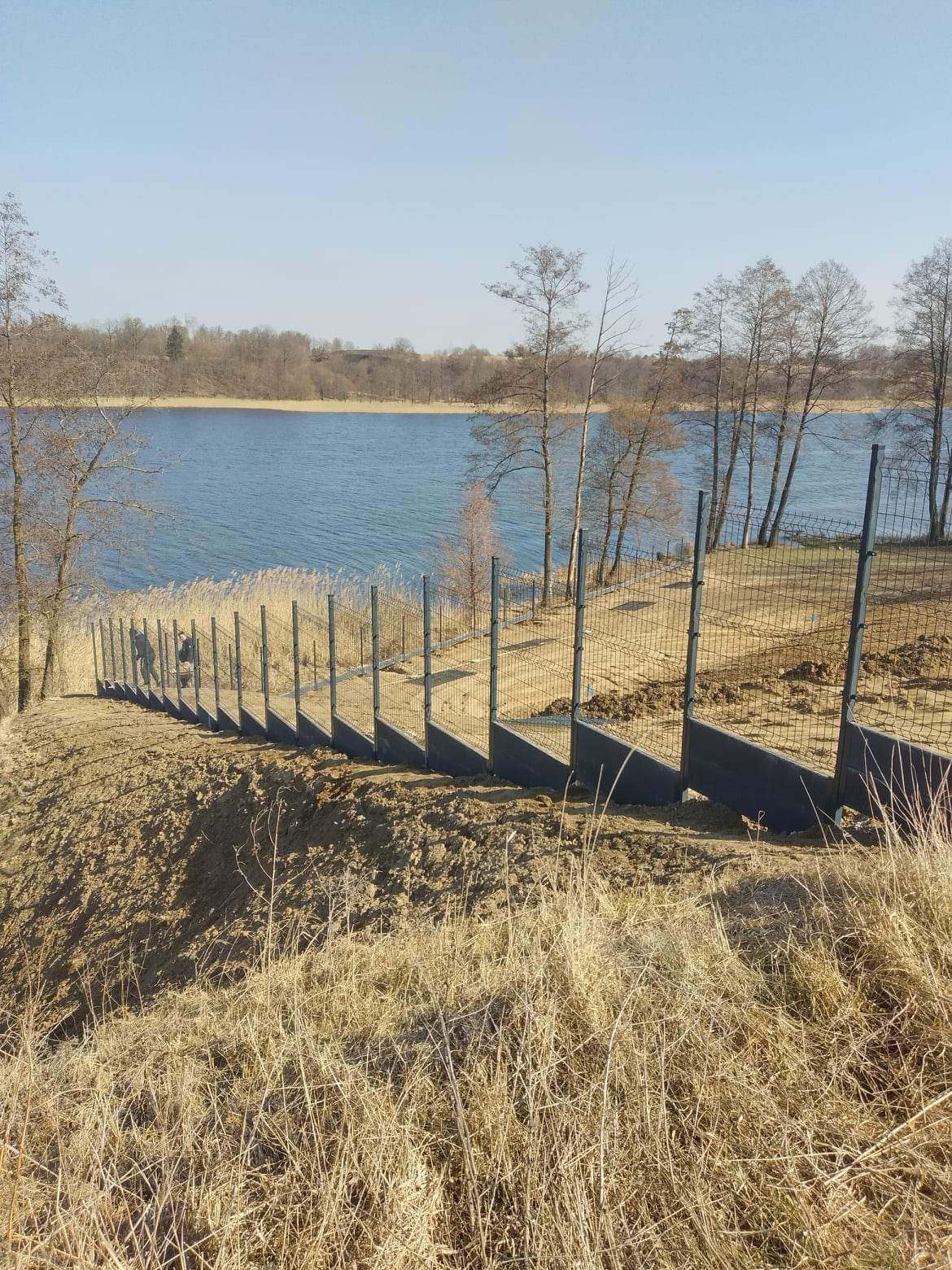 Ogrodzenie z siatki na stromej skarpie schodzącej do jeziora, widoczne słupki i podmurówka, w tle woda i drzewa bez liści.