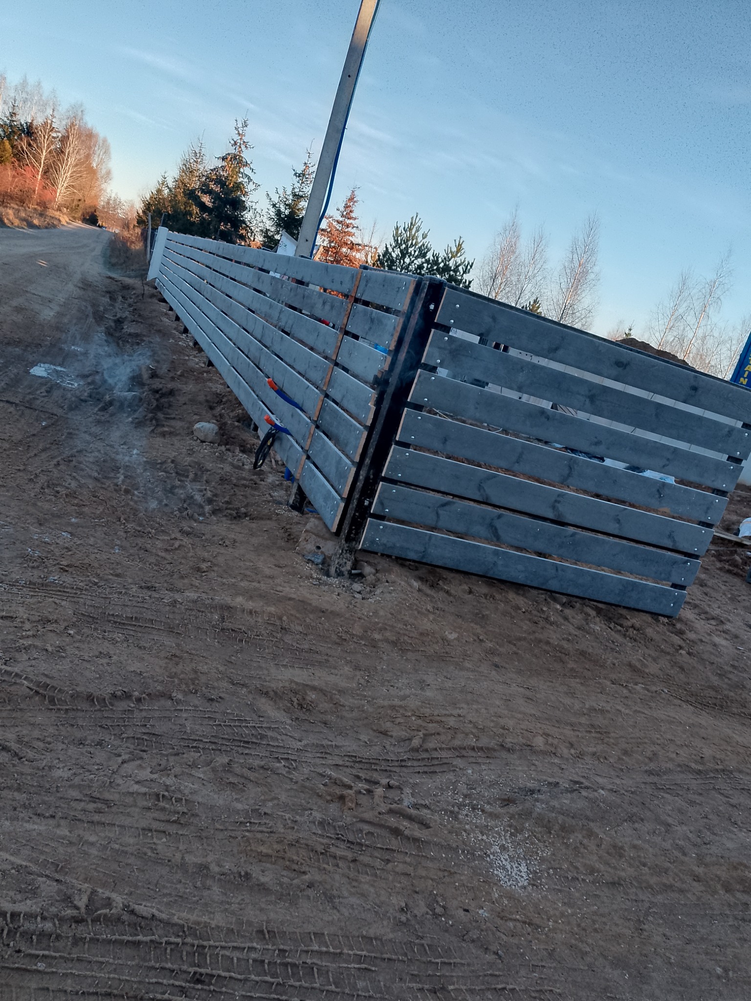 Fragment nowo postawionego, szarego ogrodzenia z poziomych desek, widoczne ślady opon na błotnistym podłożu i dym w tle, sugerujący niedawne prace ziemne.