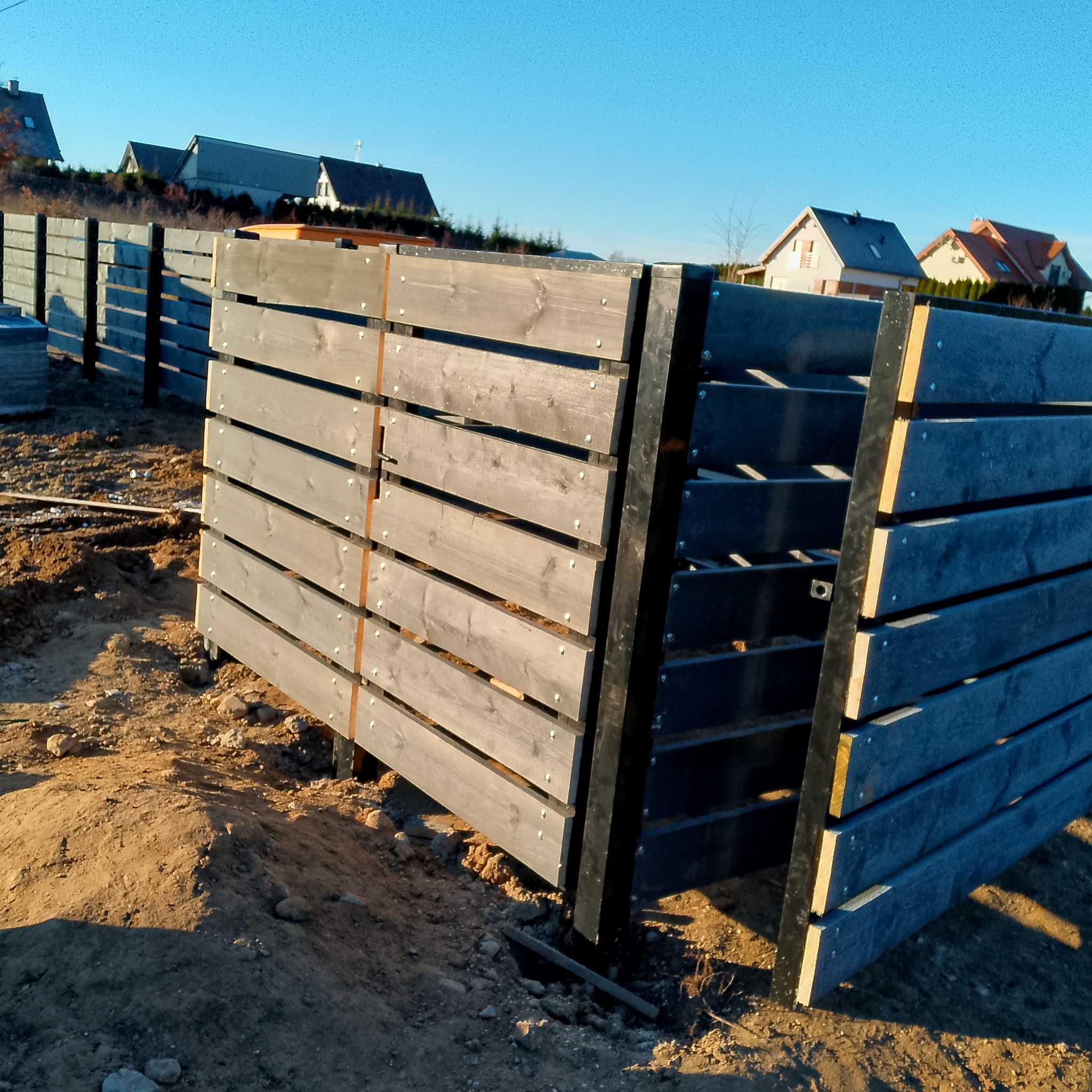 Fragment budowy ogrodzenia panelowego z drewnianymi deskami i metalowymi słupkami, widoczne przygotowane podłoże i domy w tle.