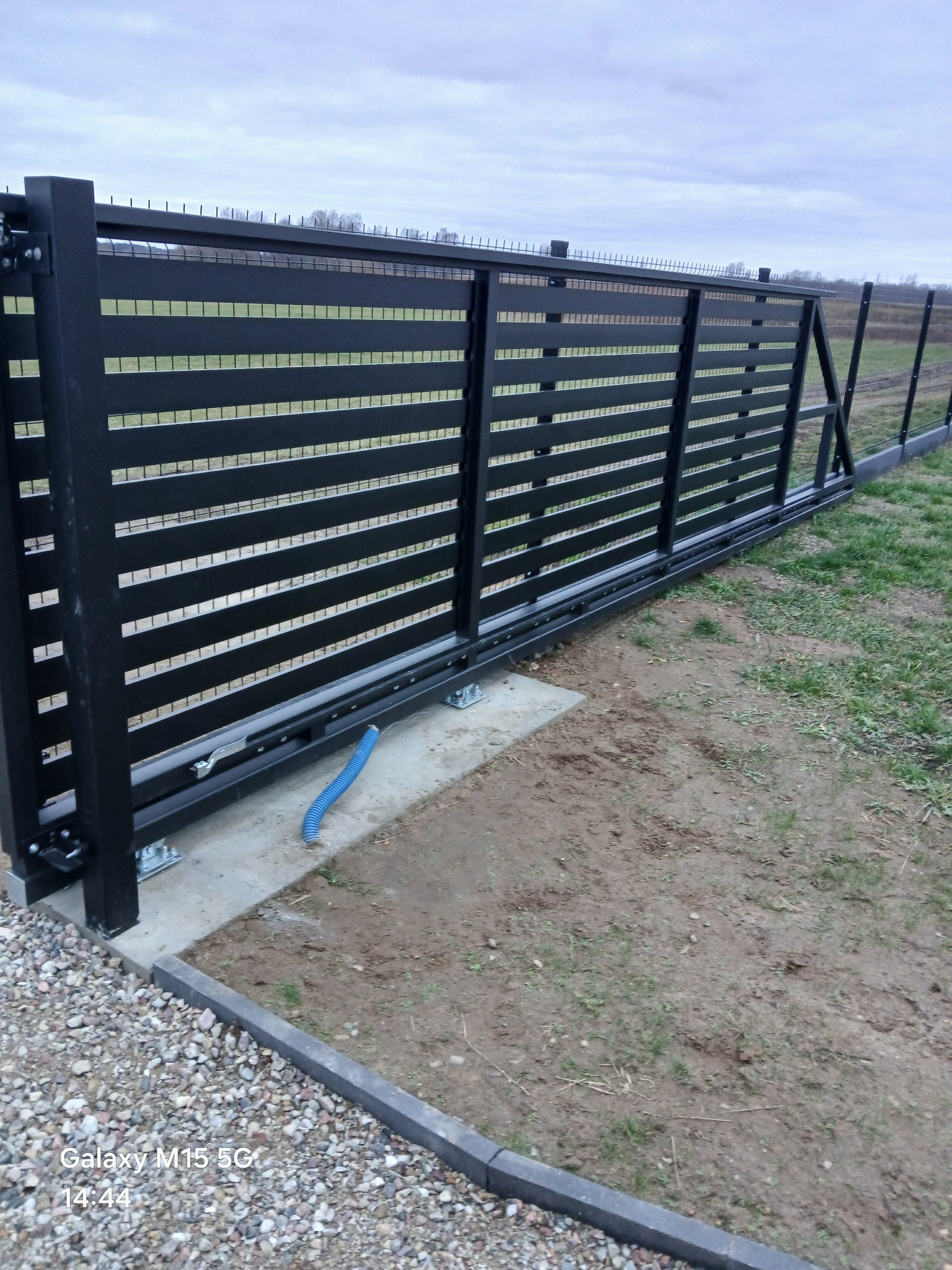 Nowoczesna, przesuwna brama w kolorze antracytu z poziomymi panelami, zamontowana na betonowej podstawie. Widoczny fragment instalacji elektrycznej i otoczenie z kamieniami.