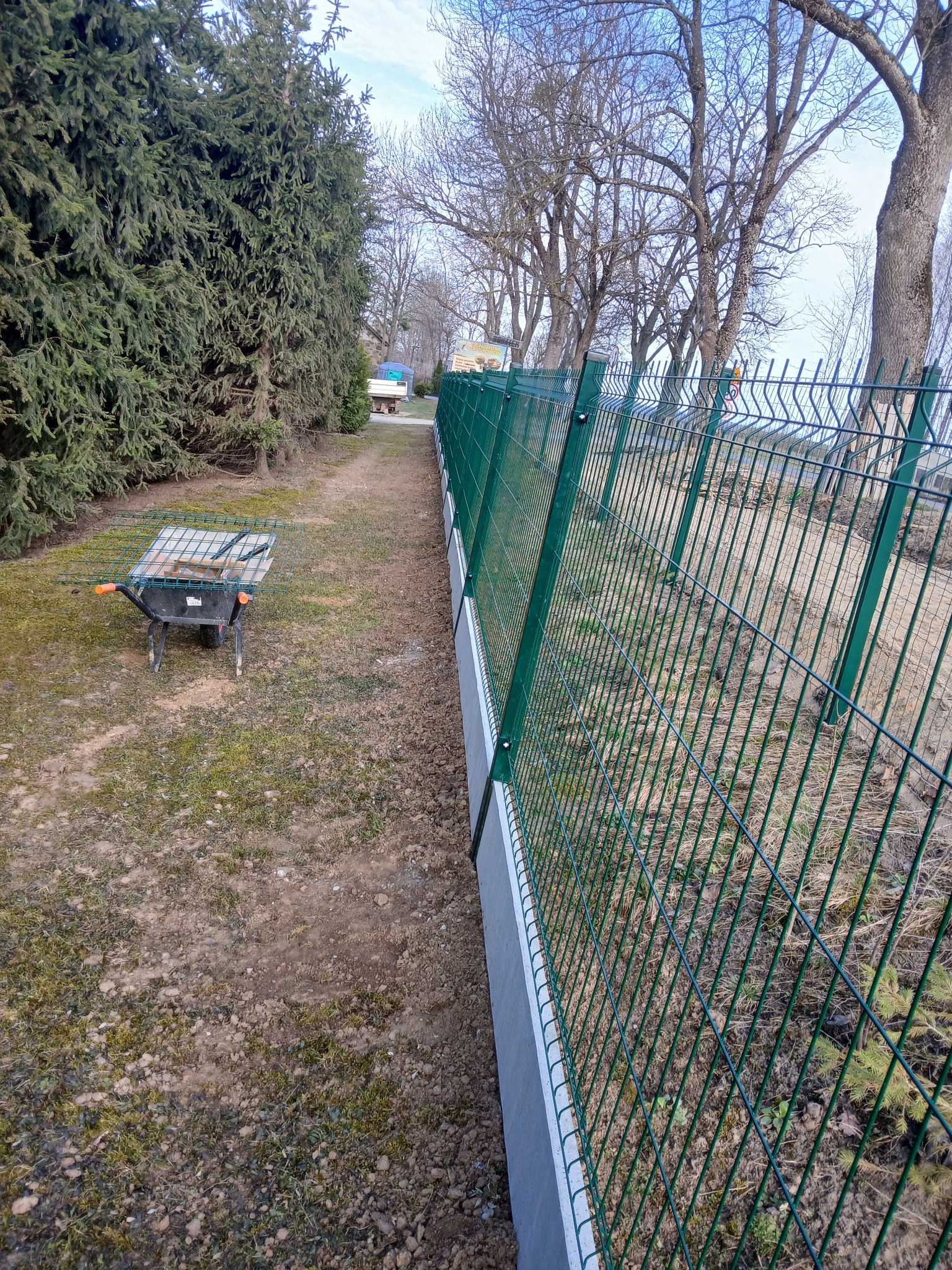 Zielone ogrodzenie panelowe na betonowej podmurówce, ciągnące się wzdłuż granicy działki z widoczną taczką i drzewami w tle. Ujęcie z perspektywy.