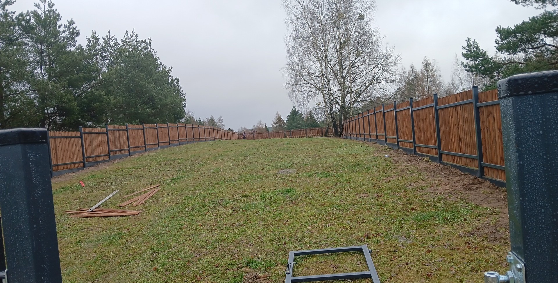 Długi, drewniany płot panelowy z metalowymi słupkami, otaczający trawnik z drzewami w tle. Widoczne elementy montażowe na pierwszym planie. Pochmurny dzień.