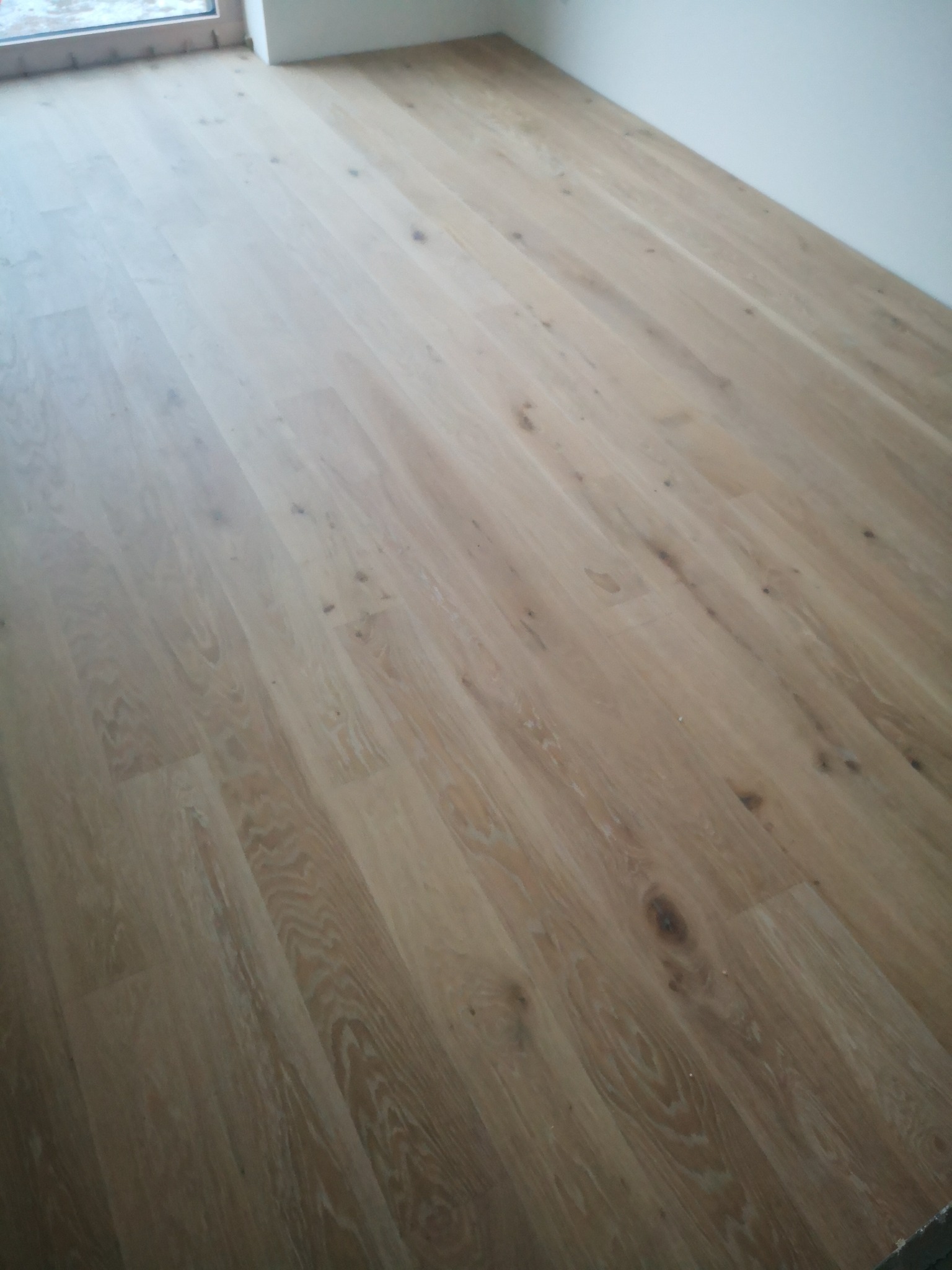 Jasne, drewniane panele podłogowe ułożone w jodełkę, widoczne sęki i naturalna struktura drewna, przy ścianie okno.