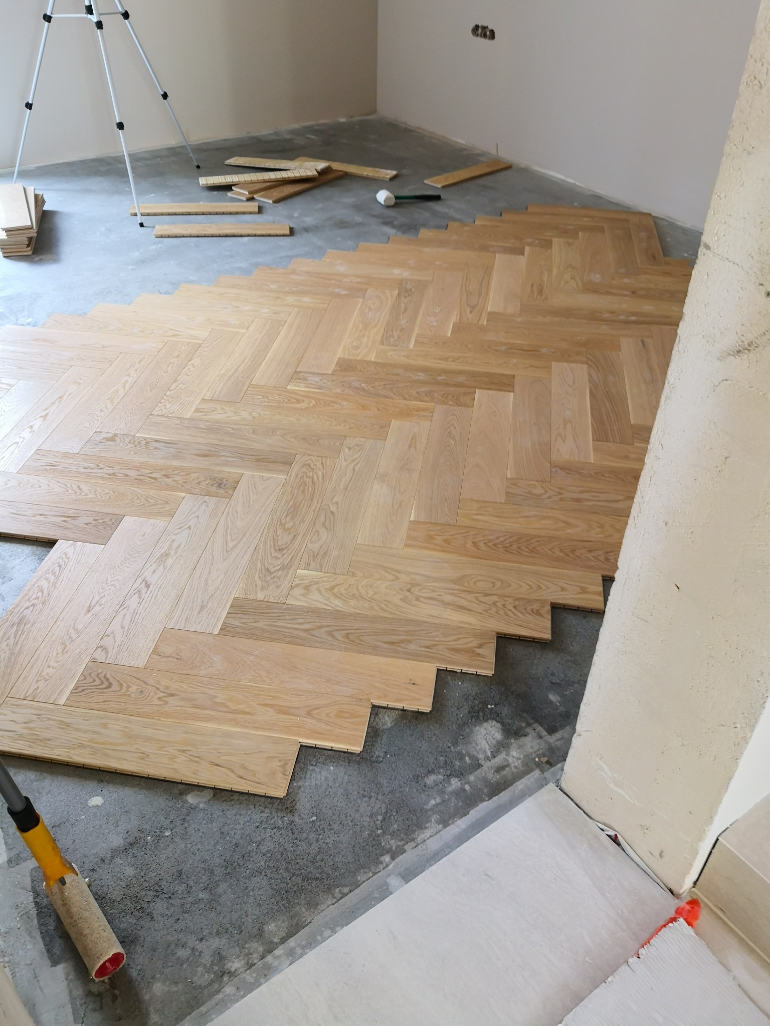 Fragment podłogi w trakcie układania parkietu w jodełkę; widoczne deski, narzędzia i surowy beton podłoża