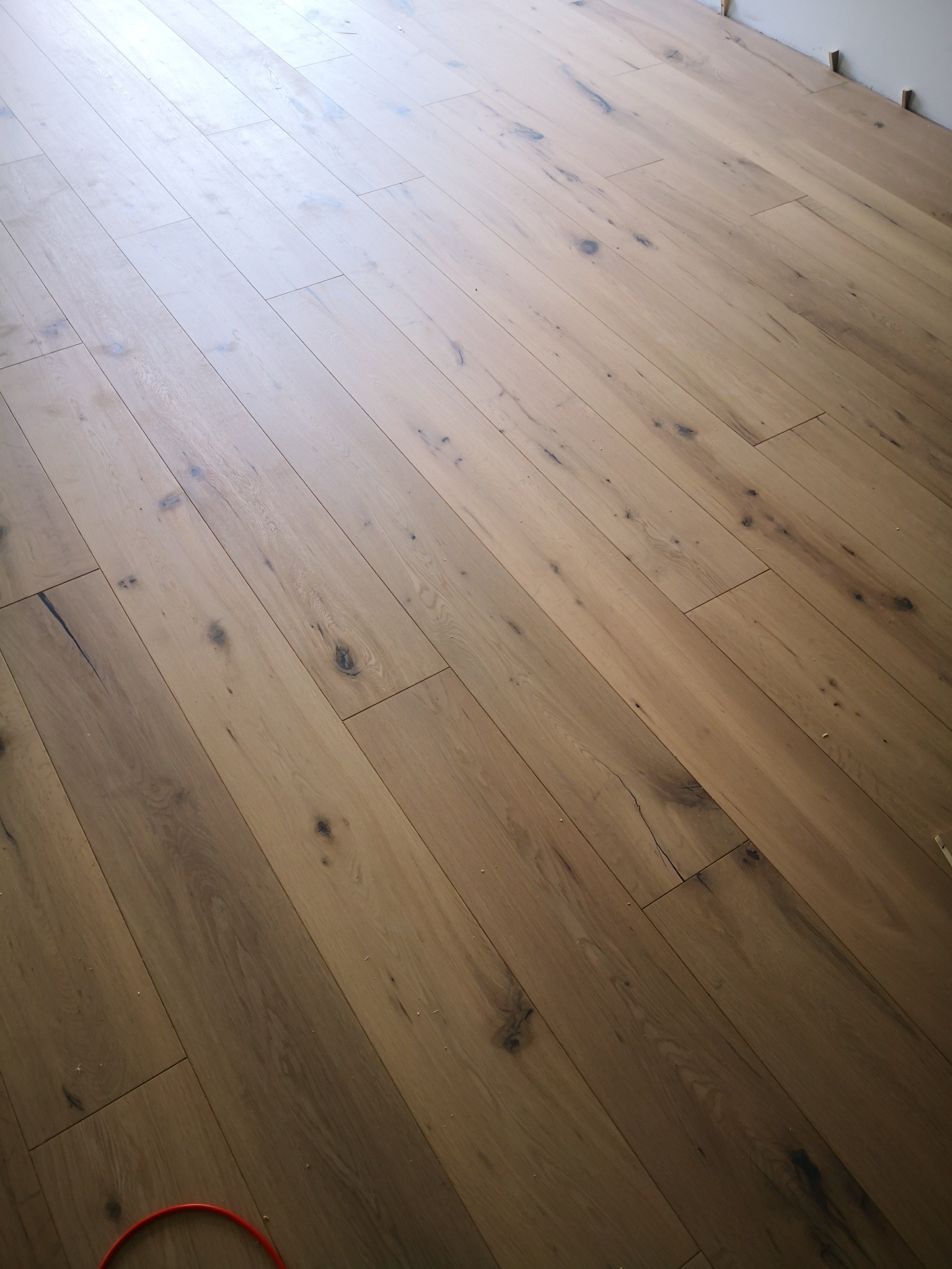 Świeżo ułożona, jasnobrązowa podłoga z desek o naturalnym wyglądzie drewna z widocznymi słojami i sękami, ułożona pod kątem, z fragmentem czerwonego kabla w rogu.