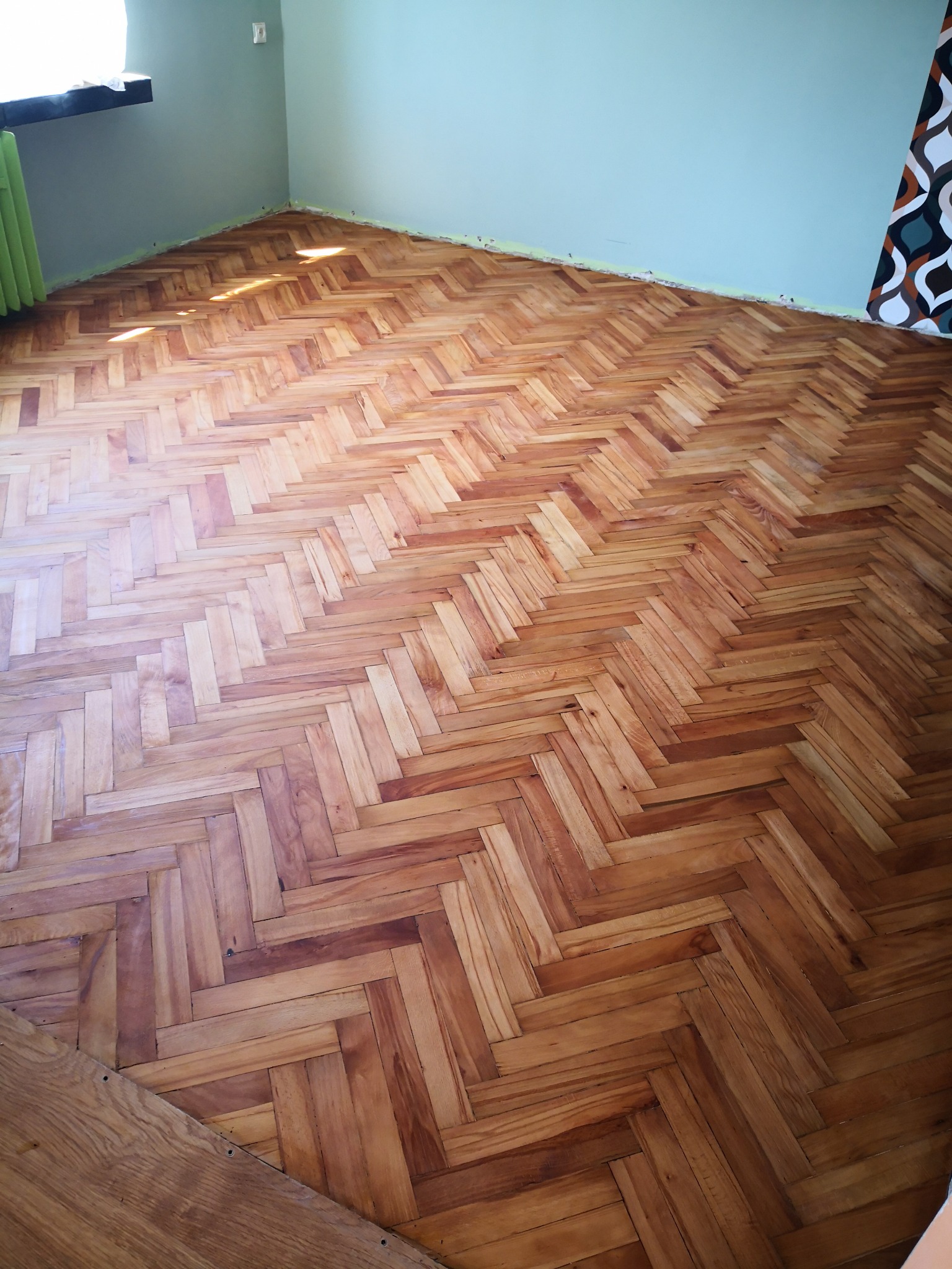 Ułożony parkiet w jodełkę w pokoju z pomalowanymi na jasnozielono ścianami, widoczne ślady po niedawnym remoncie przy listwach przypodłogowych.