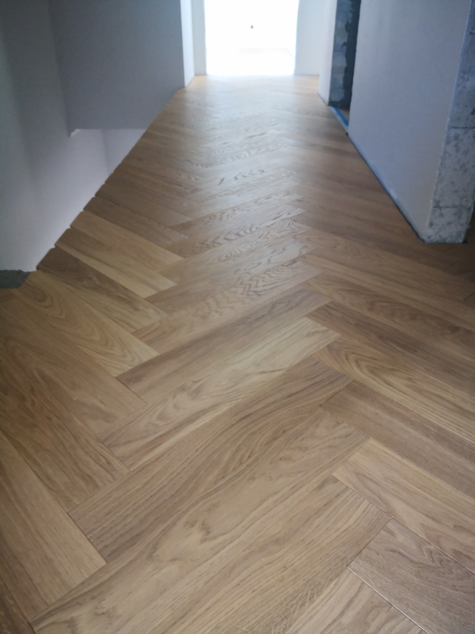 Jasnobrązowa podłoga ułożona w jodełkę w nowym budynku, widoczne surowe ściany i przejście do kolejnego pomieszczenia.