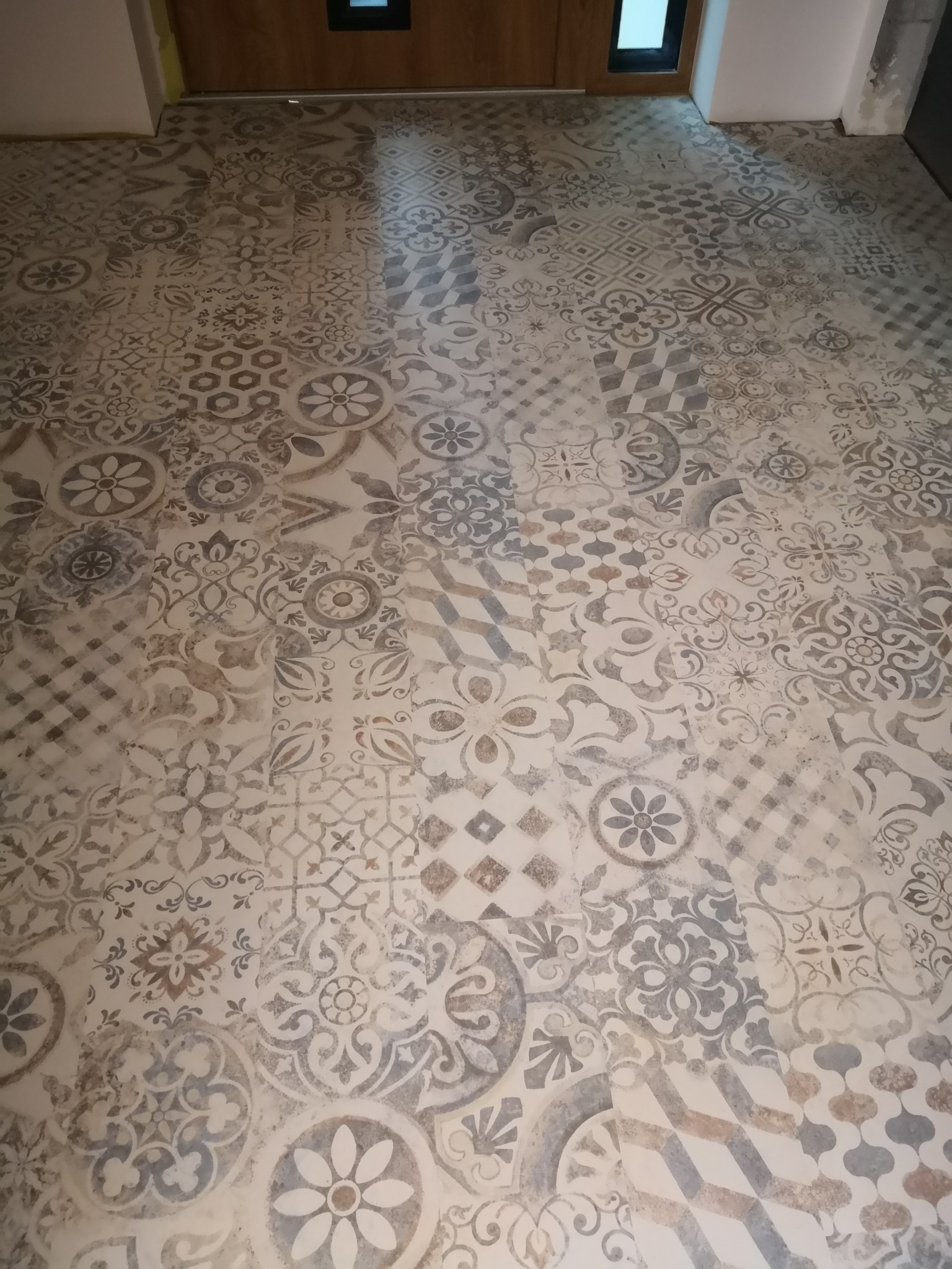 Ułożona podłoga z płytek imitujących patchwork w odcieniach beżu, szarości i bieli, z widocznymi drzwiami w tle.