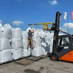 Pracownik wspina się po stosie dużych worków z zawartością, prawdopodobnie na paletach, obok wózka widłowego z żółtym osprzętem, widoczne flagi w tle.