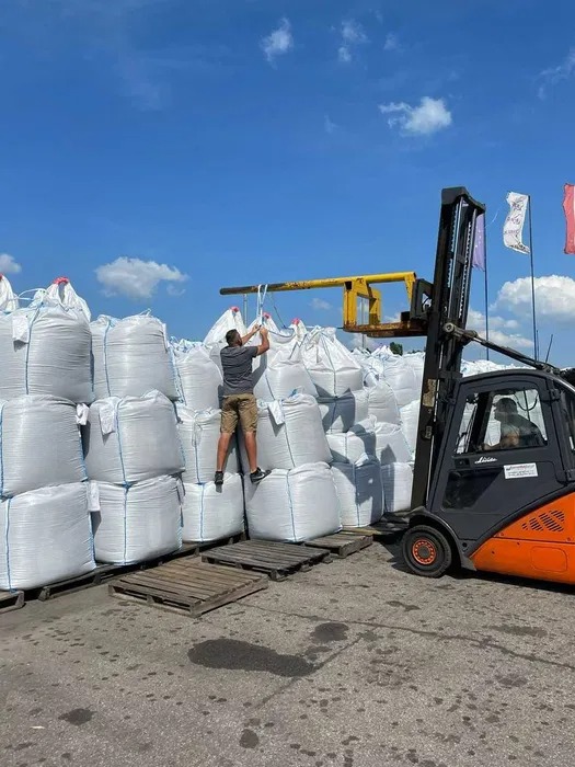 Pracownik wspina się po stosie dużych worków z zawartością, prawdopodobnie na paletach, obok wózka widłowego z żółtym osprzętem, widoczne flagi w tle.