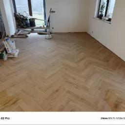 Ułożona podłoga z paneli w jodełkę, widoczne materiały budowlane, okno z rośliną w tle, ściany przygotowane do malowania.