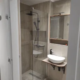 Nowoczesna łazienka z kabiną prysznicową typu walk-in z czarną armaturą i umywalką nablatową na drewnianym blacie, wykończona beżowymi płytkami.