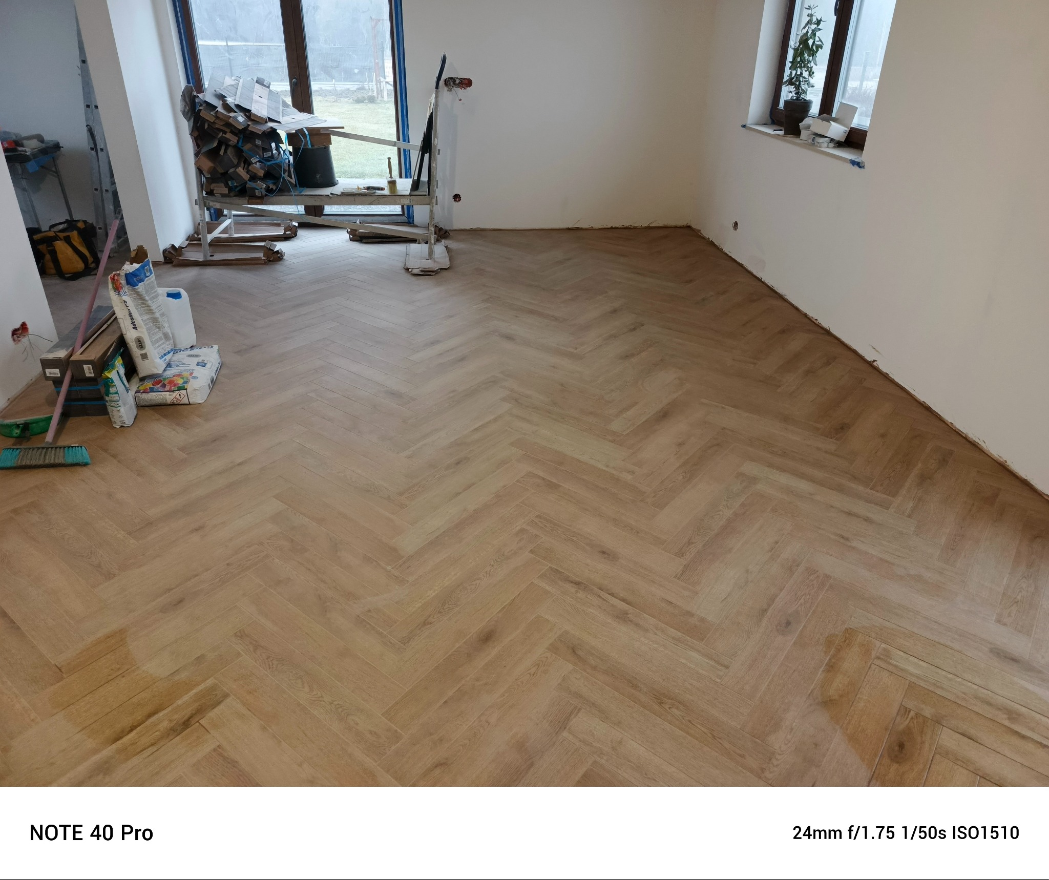 Ułożona podłoga z paneli w jodełkę, widoczne materiały budowlane, okno z rośliną w tle, ściany przygotowane do malowania.