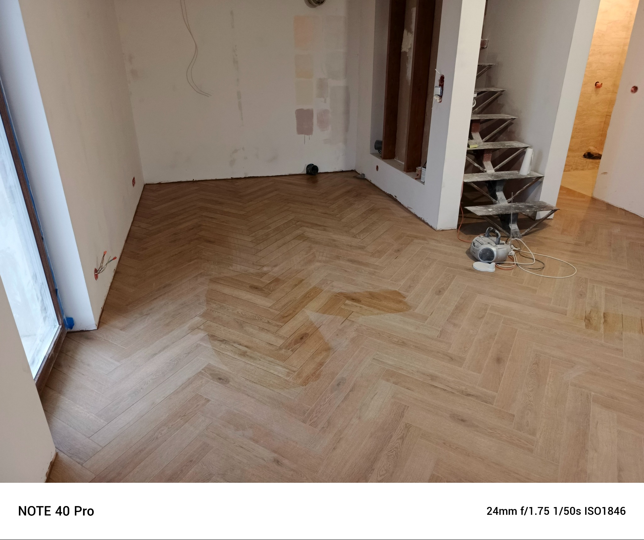 Parkiet ułożony we wzór jodełki w trakcie prac wykończeniowych, widoczne niedomalowane ściany, schody w stanie surowym i radio budowlane.