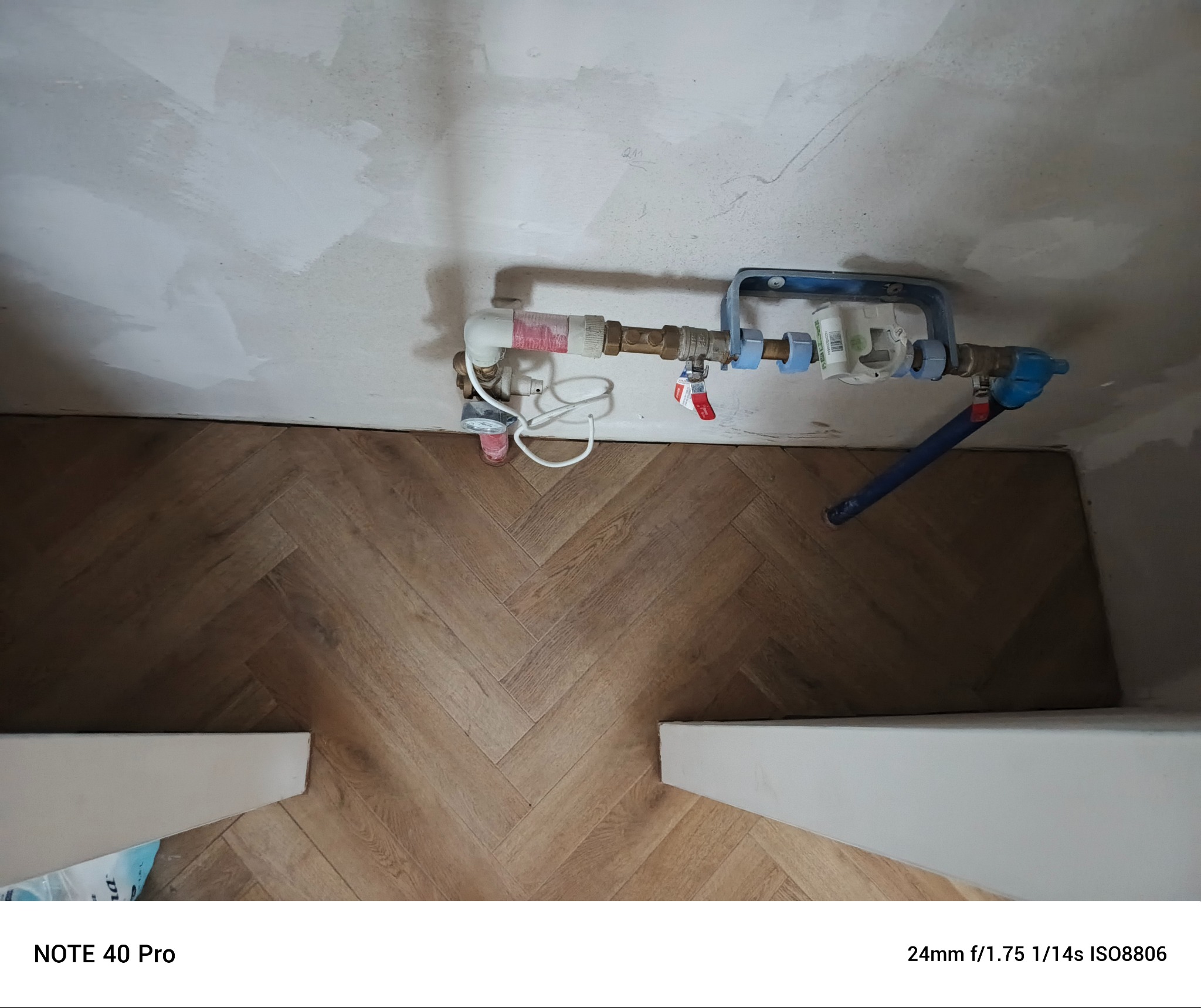 Układanie parkietu w jodełkę, widoczne rury i wodomierz przy ścianie wymagającej wykończenia.