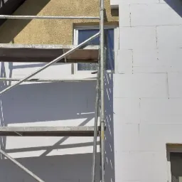 Dom w trakcie ocieplania: fragment elewacji pokryty białym styropianem, widoczne rusztowanie, okno i fragment ściany z żółtym tynkiem strukturalnym pod szczytem dachu.