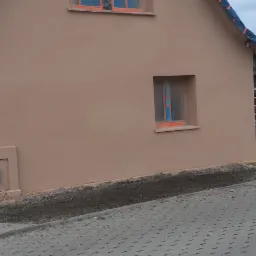 Dom jednorodzinny z nową, beżową elewacją, okna zabezpieczone folią malarską, widoczny fragment chodnika z kostki brukowej, pochmurne niebo.