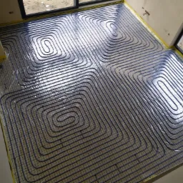 Widok z góry na ułożone, niebieskie rury ogrzewania podłogowego na folii z siatką, przyklejonej żółtą taśmą do ścian w jasnym pomieszczeniu z oknami.