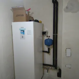 Biała pompa ciepła Panasonic Aquarea Smart Cloud zamontowana w pomieszczeniu z widocznymi rurami w izolacji termicznej, niebieskim zbiornikiem wyrównawczym i surowymi ścianami.