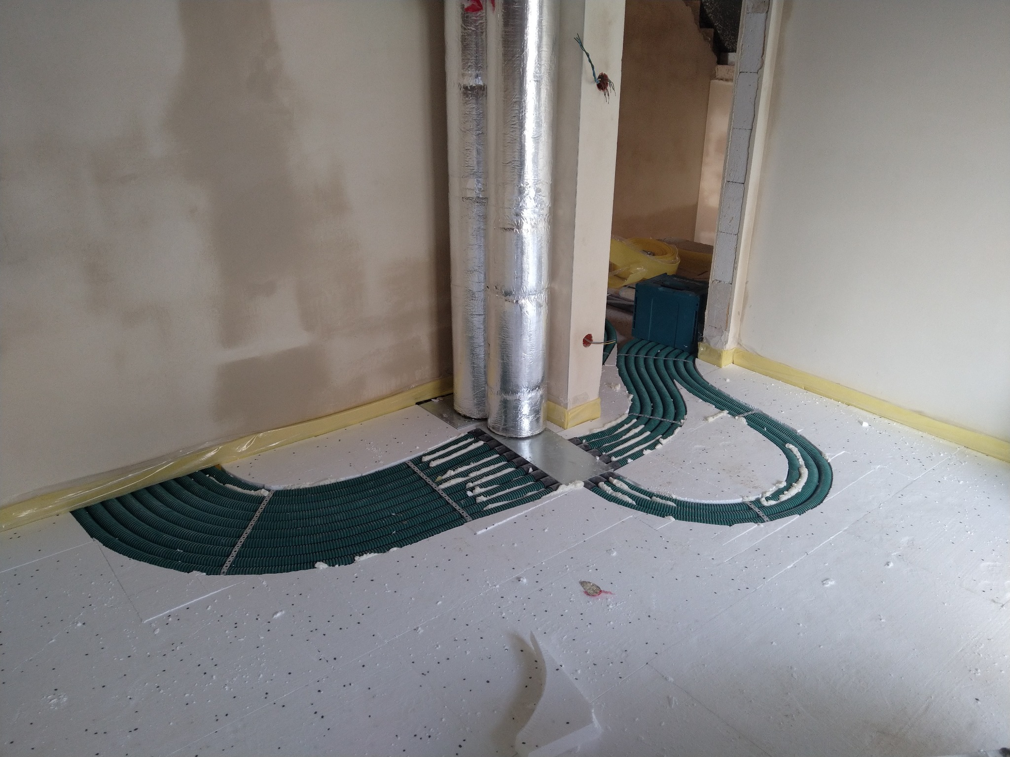 Instalacja ogrzewania podłogowego w trakcie montażu: zielone rury ułożone na białym styropianie z czarnymi punktami, dwa srebrne kominy wentylacyjne, ściany w surowym stanie, folia ochronna przy...