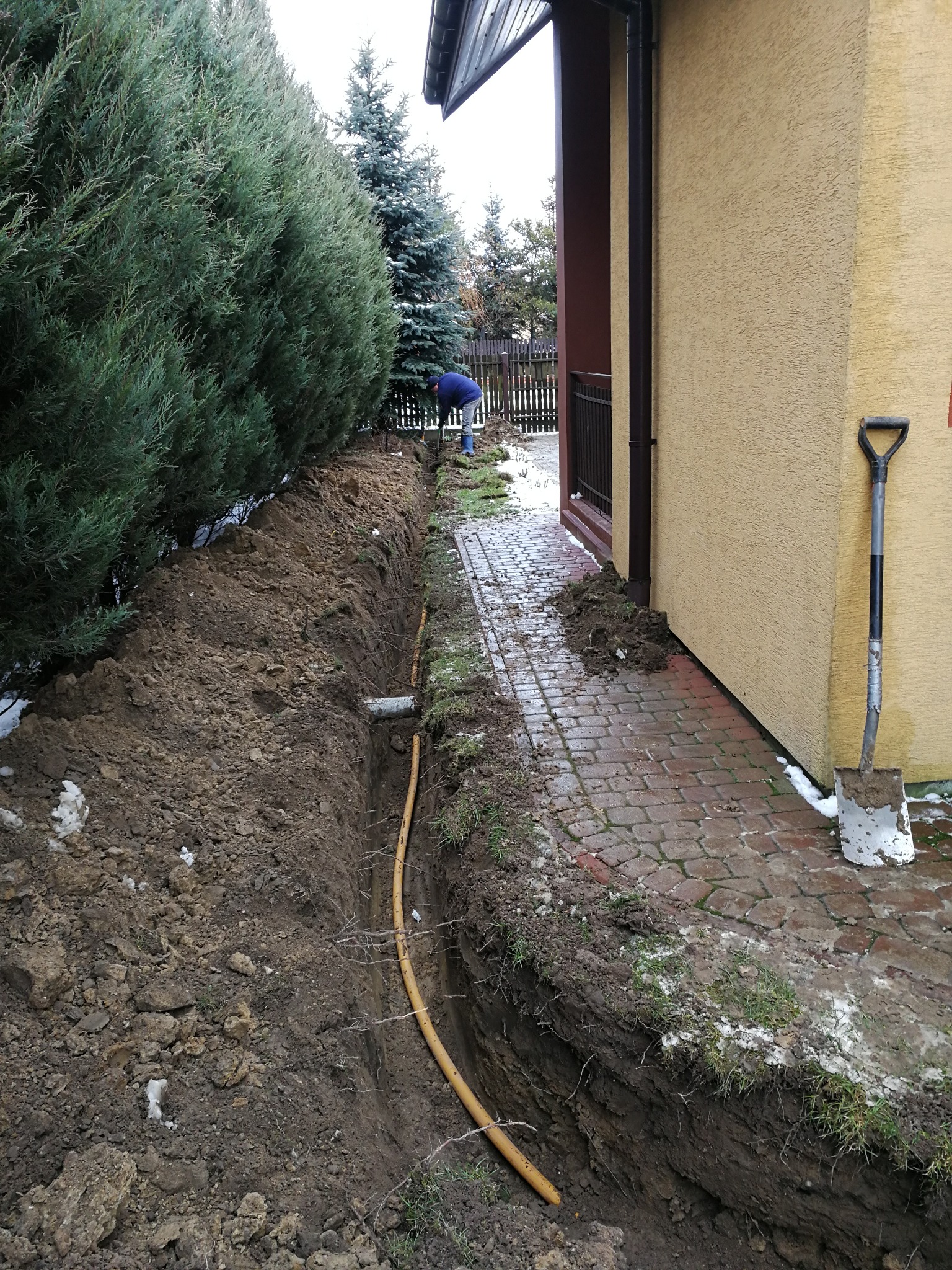 Wykop pod żółty przewód gazowy biegnący wzdłuż posesji, widoczna łopata oparta o ścianę budynku i pracownik w oddali.