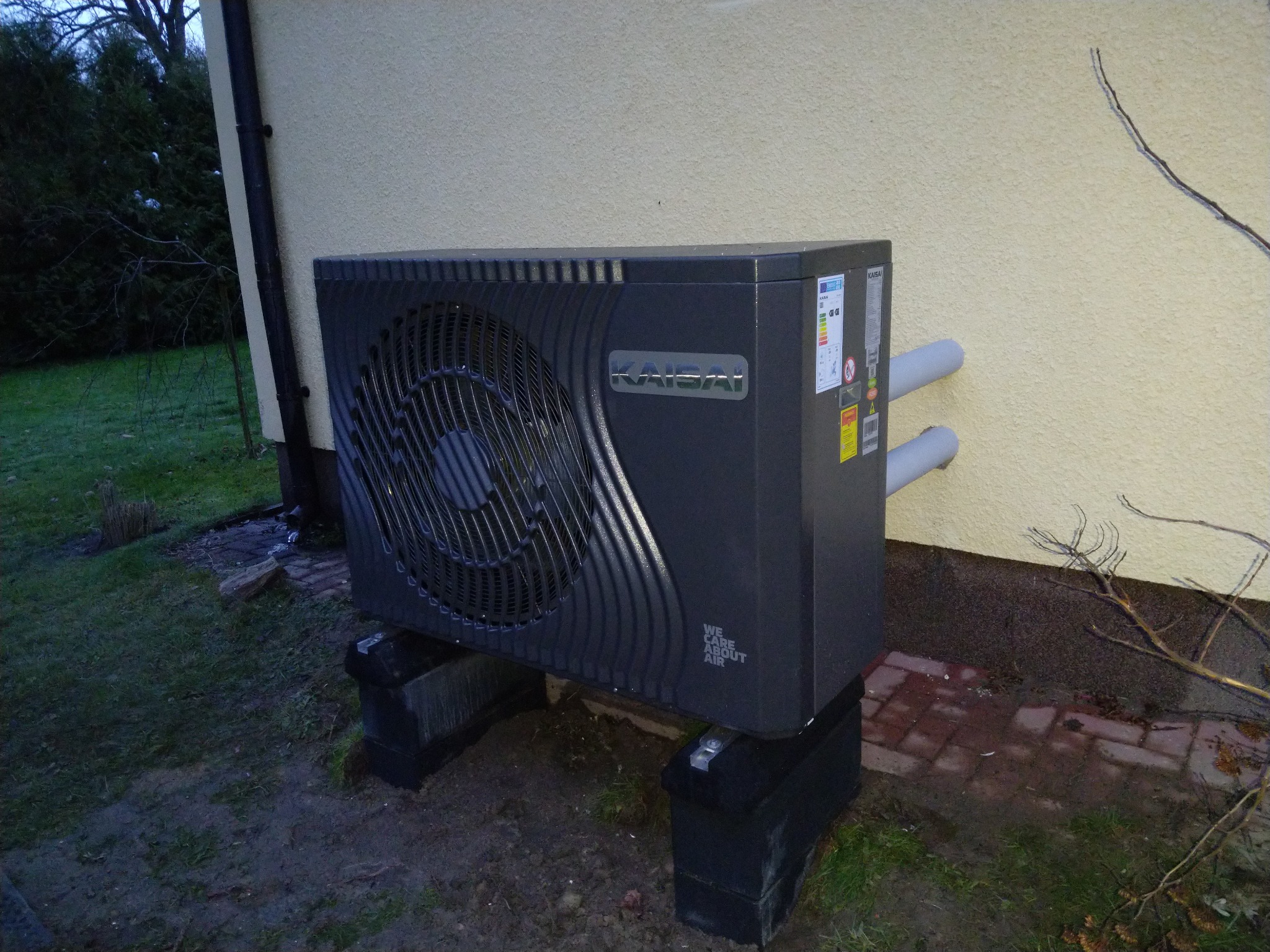 Zewnętrzna jednostka pompy ciepła marki Kaisai, zamontowana na betonowych blokach przy ścianie budynku o beżowej elewacji. Widoczne rury przyłączeniowe i naklejki informacyjne na obudowie.