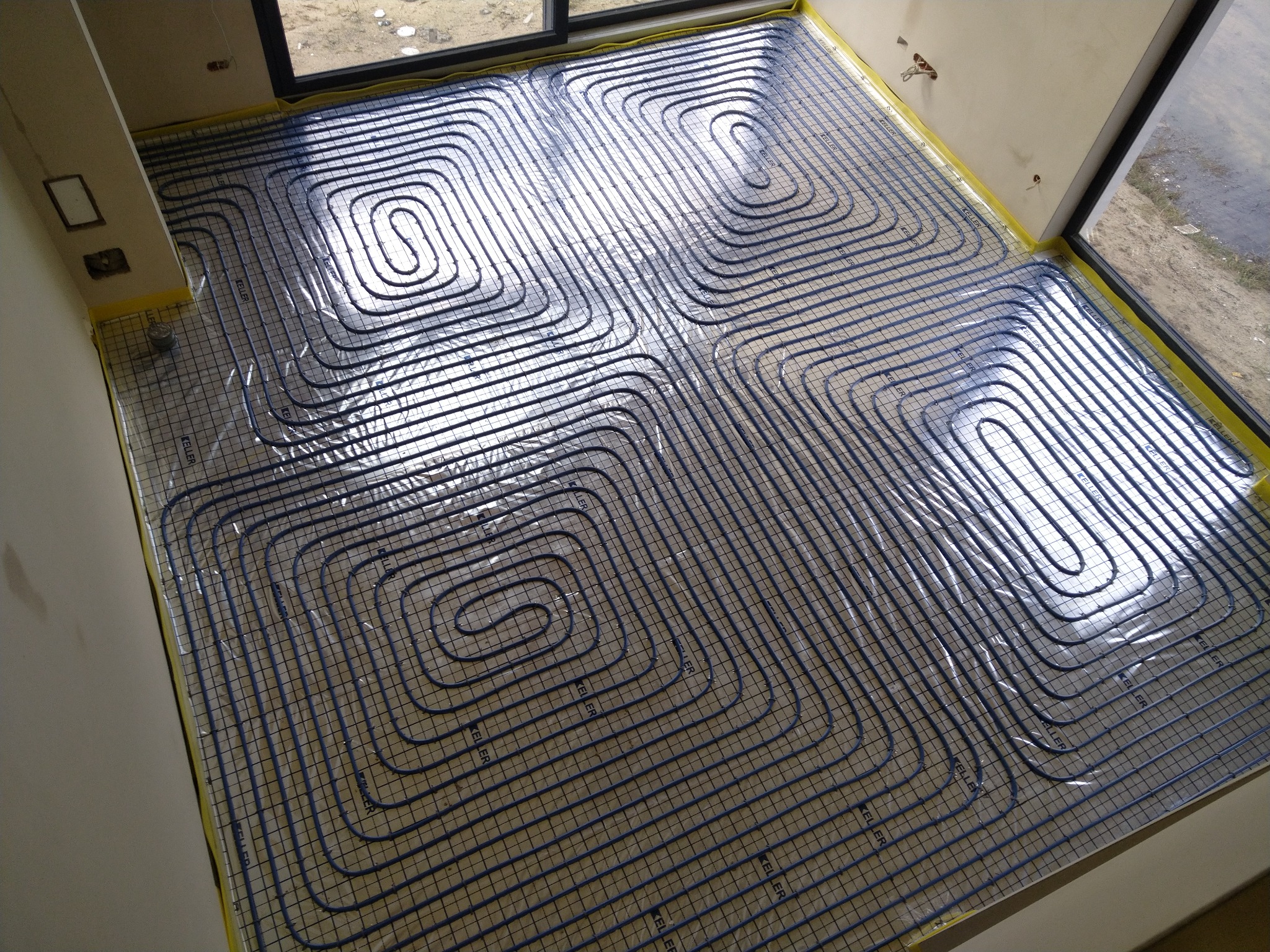 Widok z góry na ułożone, niebieskie rury ogrzewania podłogowego na folii z siatką, przyklejonej żółtą taśmą do ścian w jasnym pomieszczeniu z oknami.