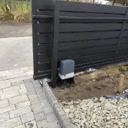 Czarna, przesuwna brama wjazdowa z automatycznym napędem, widoczny mechanizm i fundament w trakcie prac wykończeniowych z kamieniami.