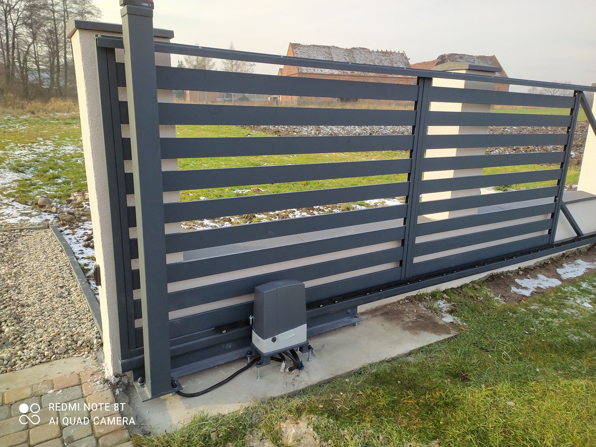 Nowoczesna, przesuwna brama w kolorze grafitowym z poziomymi panelami, zamontowana na betonowym fundamencie z widocznym automatem do bram, trawnik z resztkami śniegu w tle.